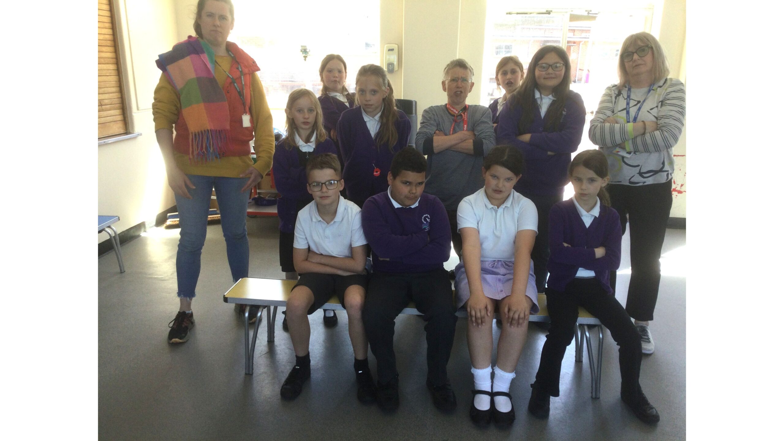 A row of four school children seated on a bench behind them another row of children, flanked by two adults with a third in the middle with arms firmly crossed