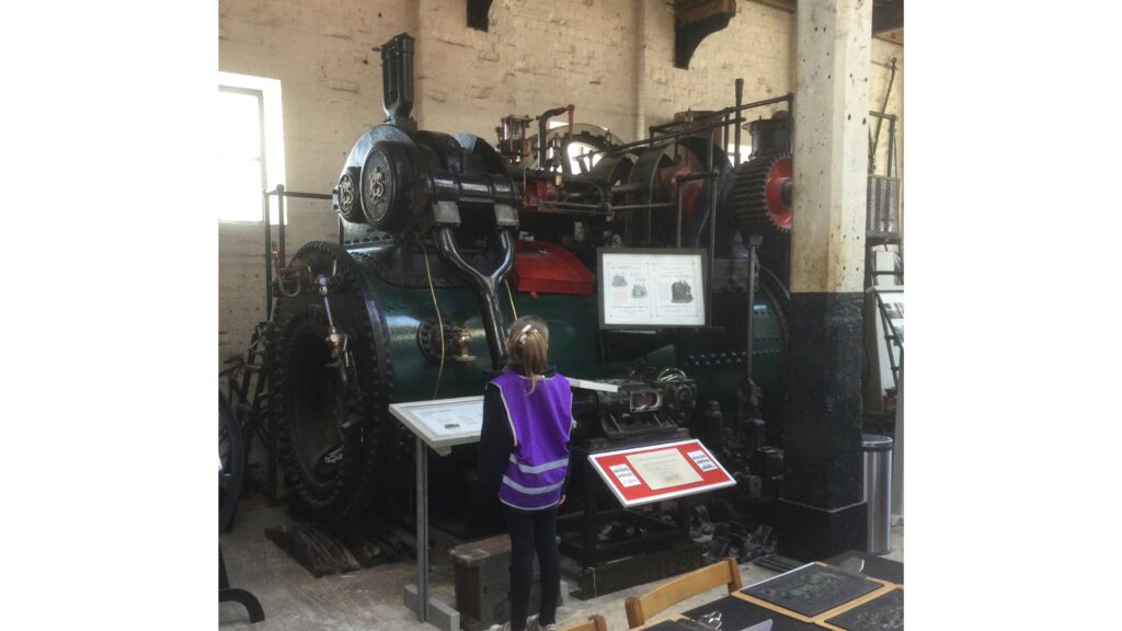 A child in a purple tabard gazes up at a green and red steam engine. There are notice boards in front, and to the side and a large concrete column, black with off-white above, to her right
