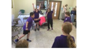 Children enact a scene from a football match as a member of staff stands in the background, looking on