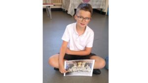 A schoolboy in black shorts and white T-shirt, with short hair and brown-rimmed glasses sits crossed legged on the floor holding a photograph of a football team in front of him. His left arm is cross over, copying the stance of one of the players in the picture