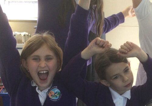 Four school girls and a member of staff, three seated with two standing behind, with arms in the air and looking joyful