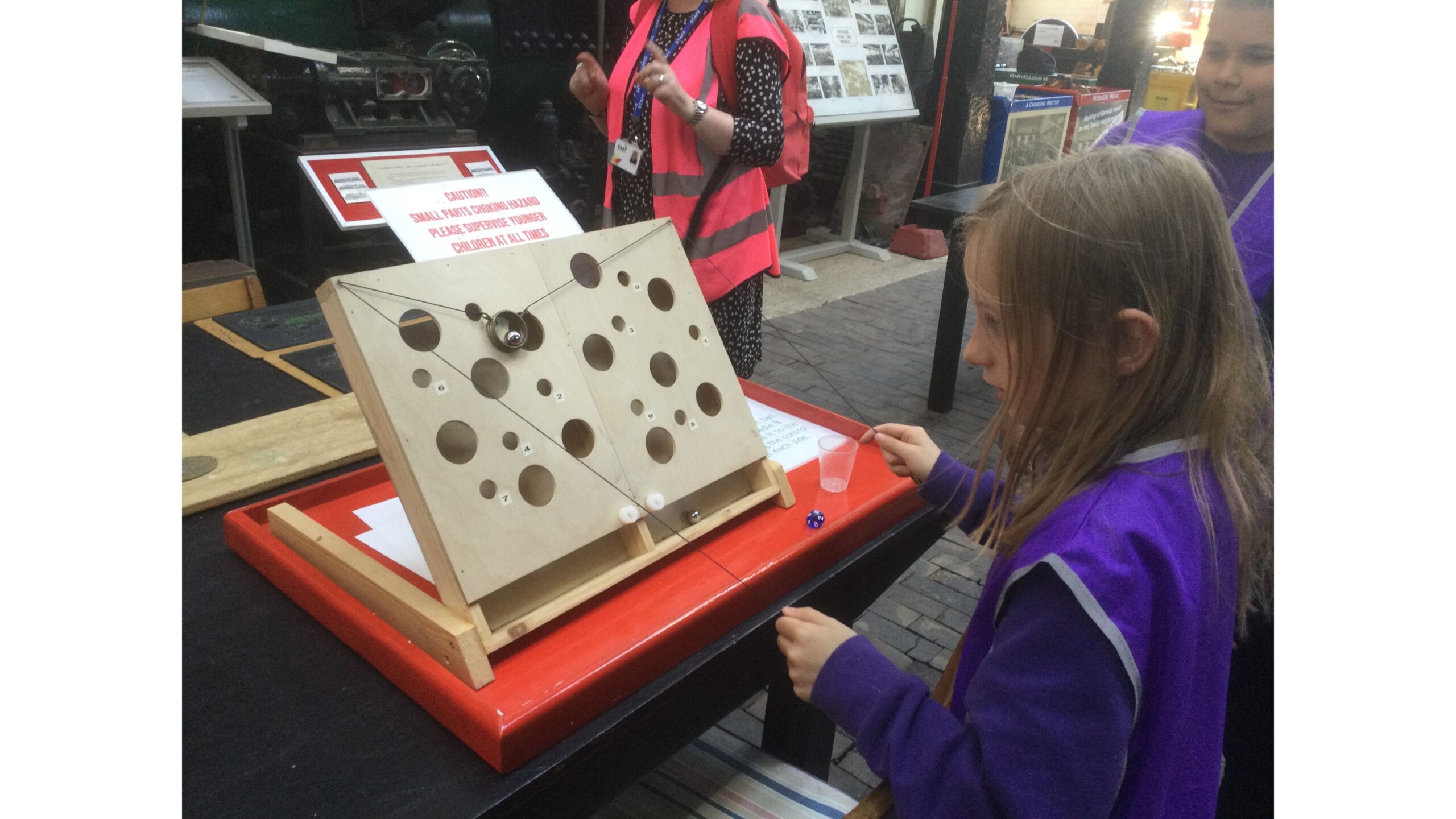 A child with long hair and wearing a purple tabard is playing a game of dexterity in the museum. She has to use two pieces of thread to move a ring and position a ball over a hole so that it falls in. Beside her is a class mate and there is an adult in a pink tabard in the back ground