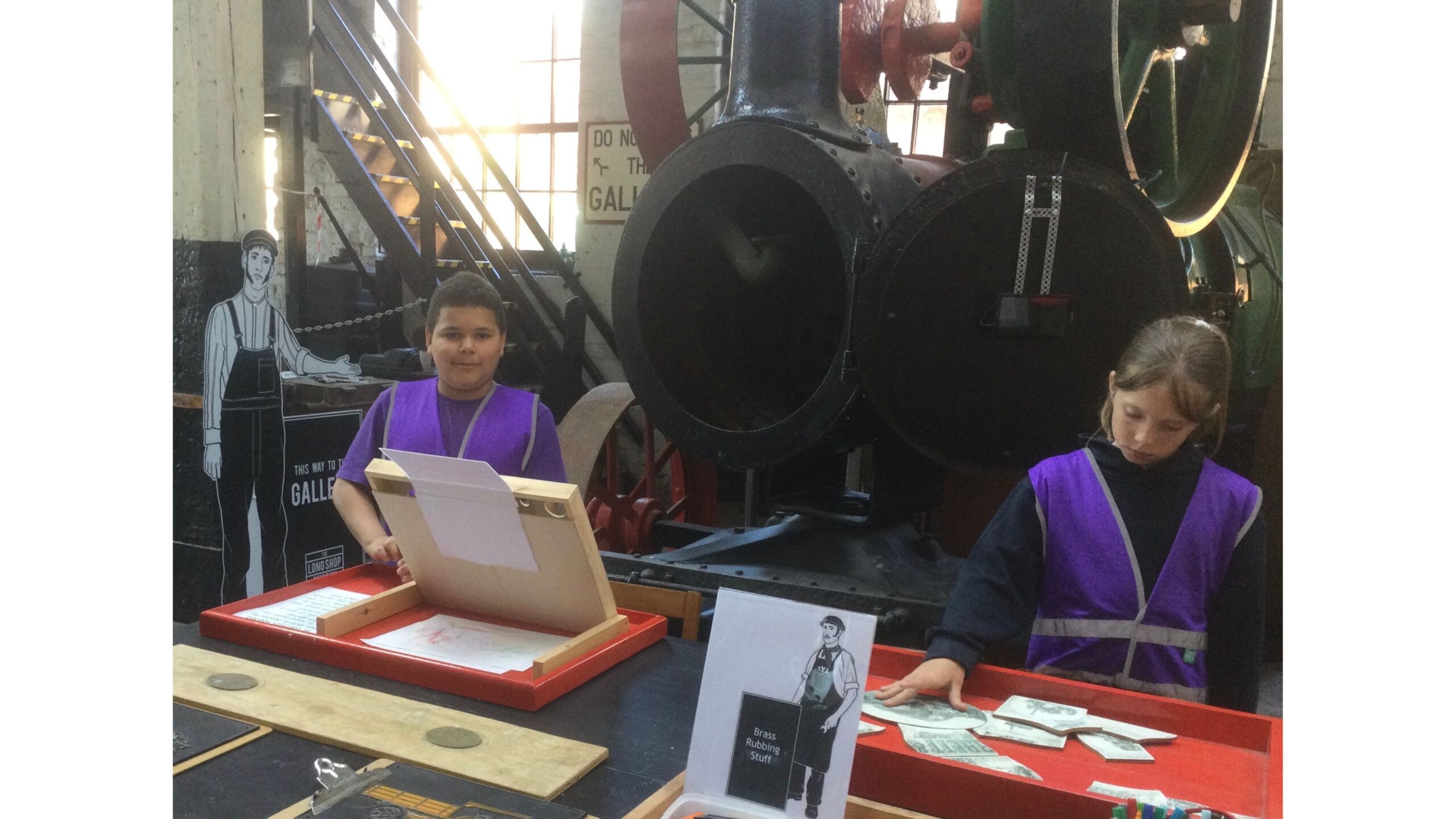 Two school children, each wearing a purple tabard over their uniform, amuse themselves with items on a table in front of them. Behind is a steam engine with the funnel door open and to the left of picture a steep flight of wooden stairs lead up in front of a large window