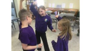 Three school children stand in large space with grey tables and chairs in the background. The children are dressed in purple tops and black trousers and they have their arms raised and clearly laughing