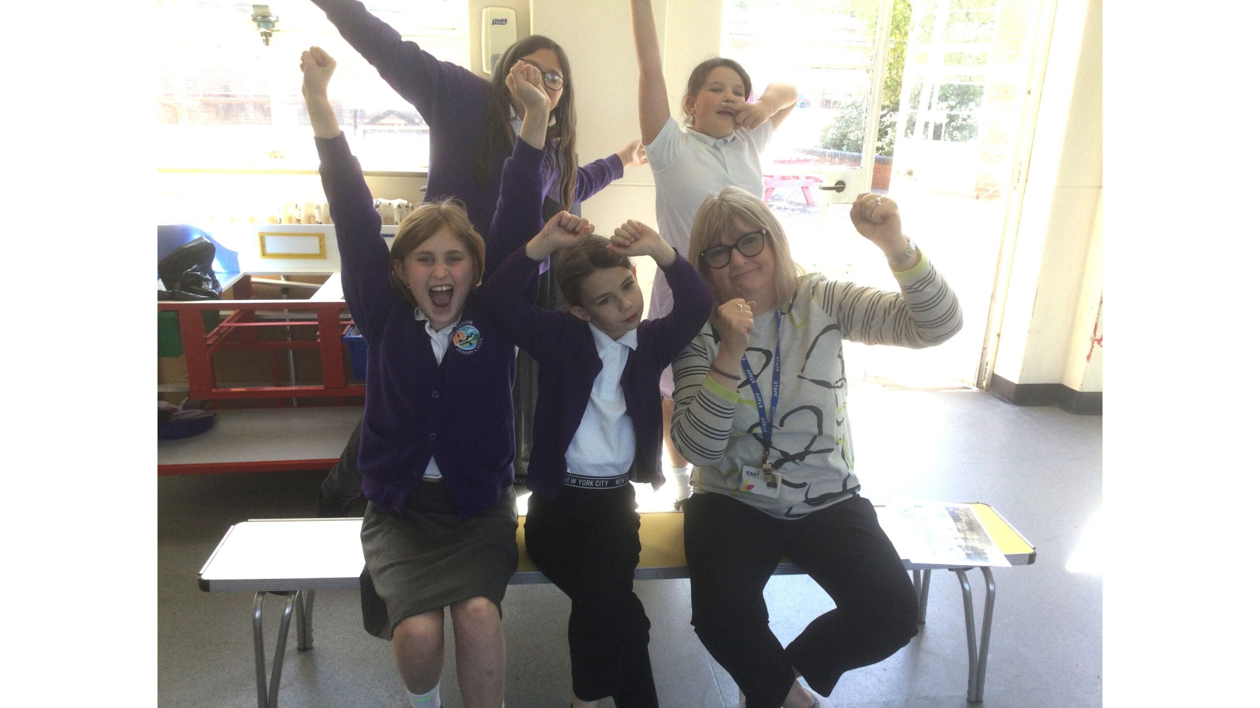 Seated on a school bench are two children and an adult; behind them are two more children. They all have their arms in the air, as if celebrating something