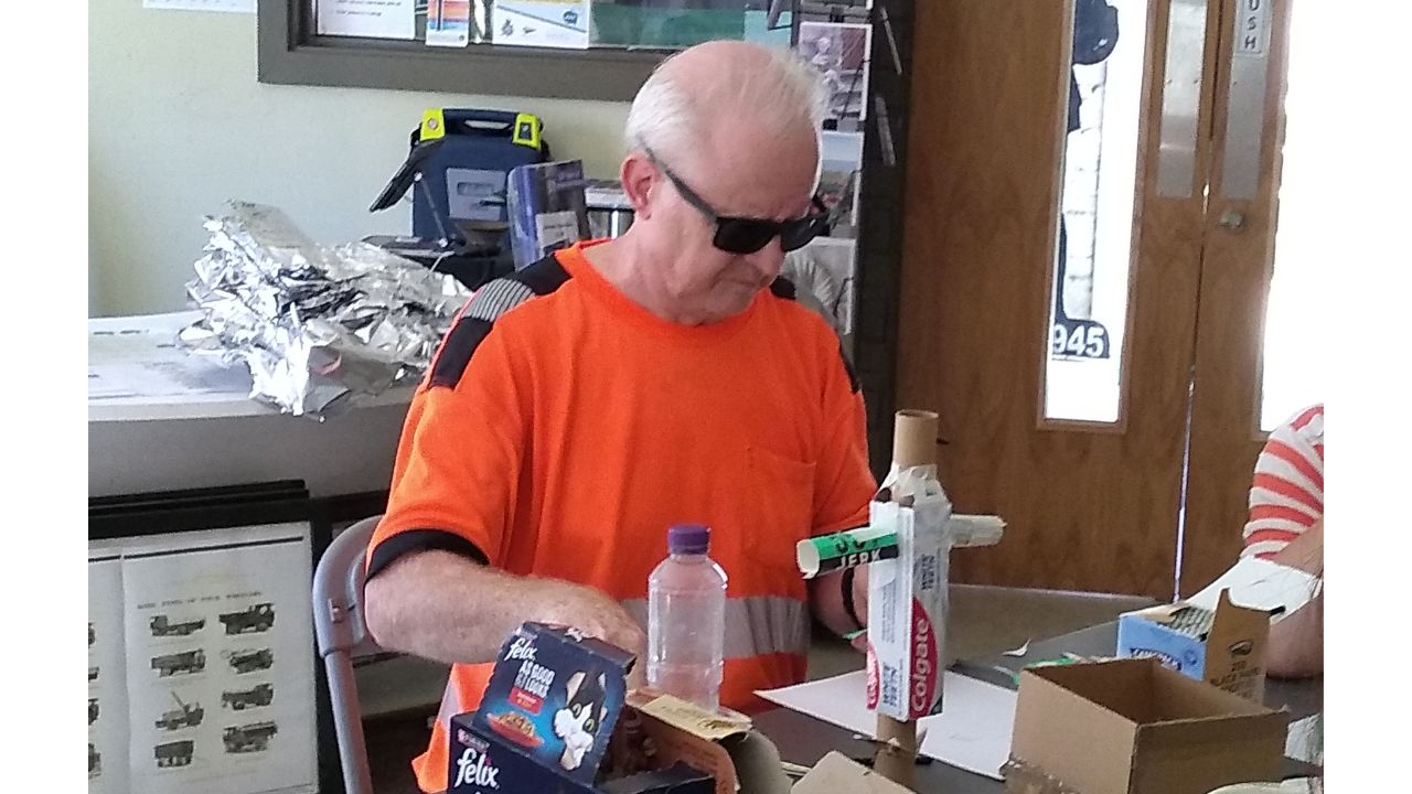 A gentleman in an orange top and wearing sunglasses has a selection of small cardboard boxes and other craft materials around him and appears to be fixing a toothpaste tube to a circular tube with small tubes either side