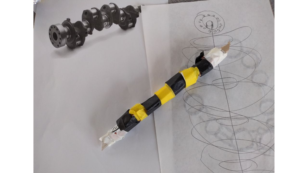 A monochrome print of an engineered part with moving elements, a drawing of same and a yellow and black cardboard model