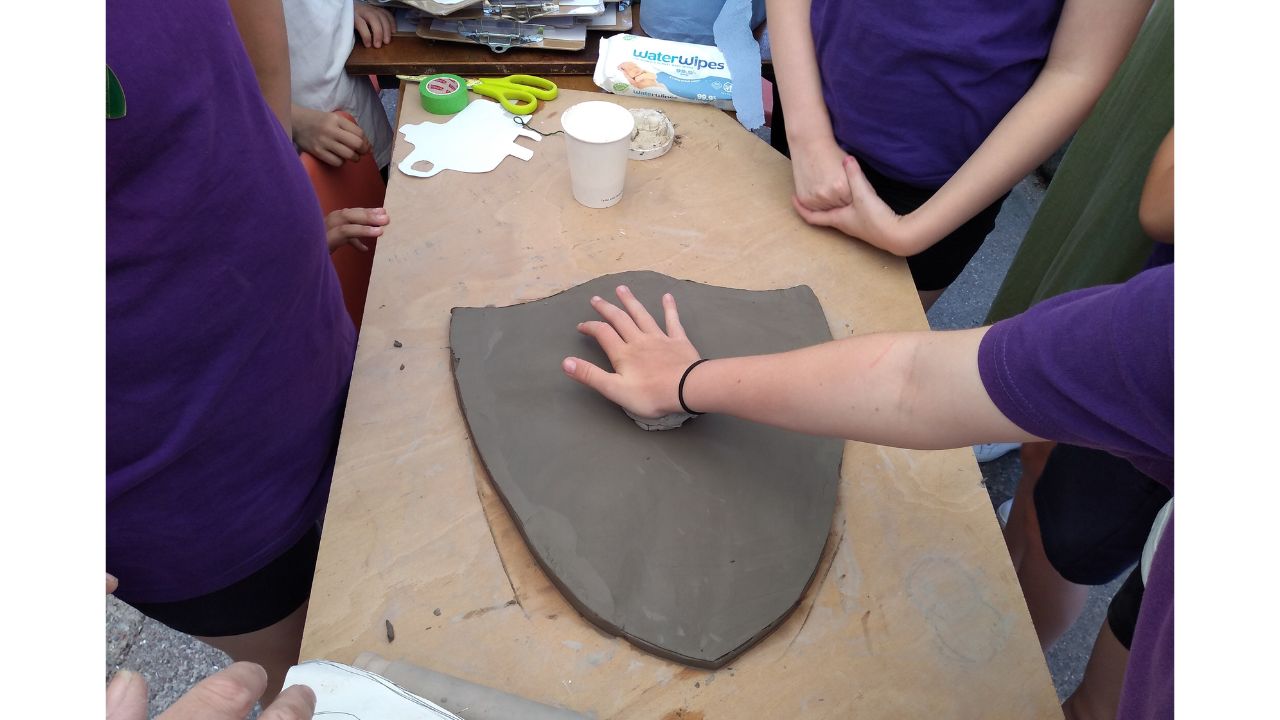 Overhead shot of child's arm, pressing something into a large shield shaped piece of clay