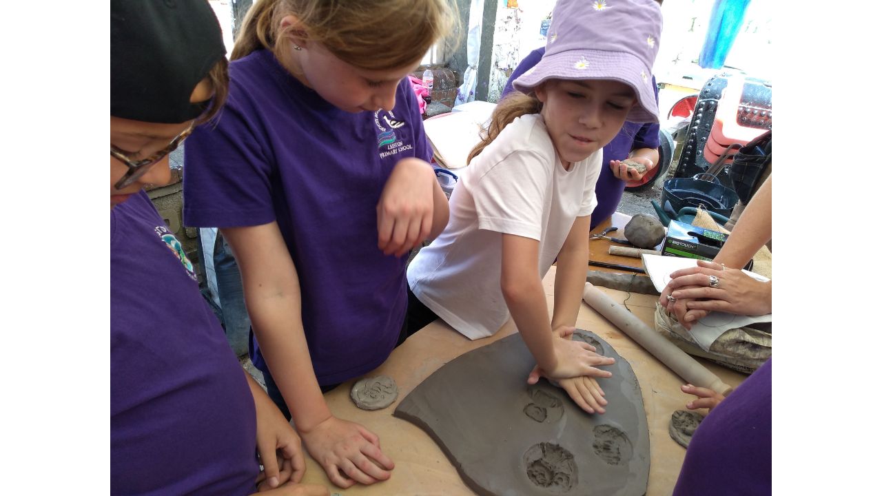 A child in white T shirt and lilac and white spotted sun hat pressed her mould firmly into the clay as other children look on