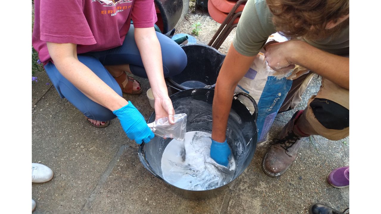 Adult hands reach into a large black bucket to mix the plaster and add a packet of metal powder