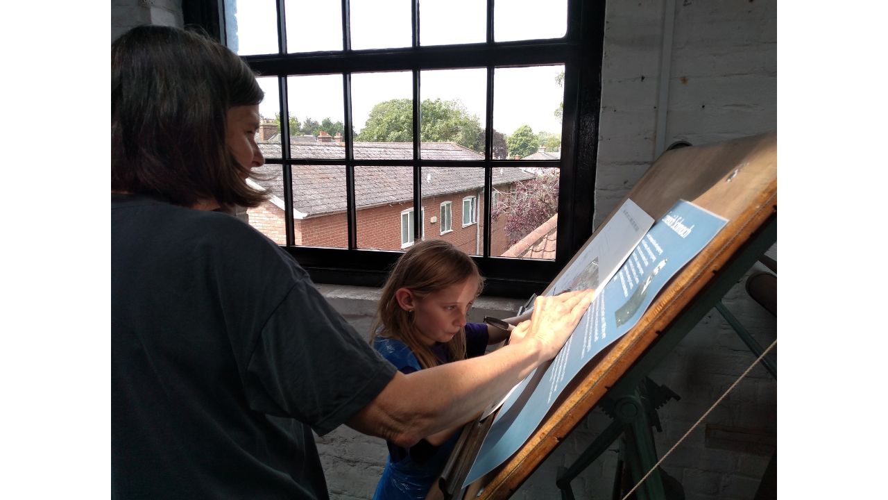 A child stands before a drawing board, intent on drawing something, as an adult looks on. Behind is a large multi-paned window with a view out over a row of houses