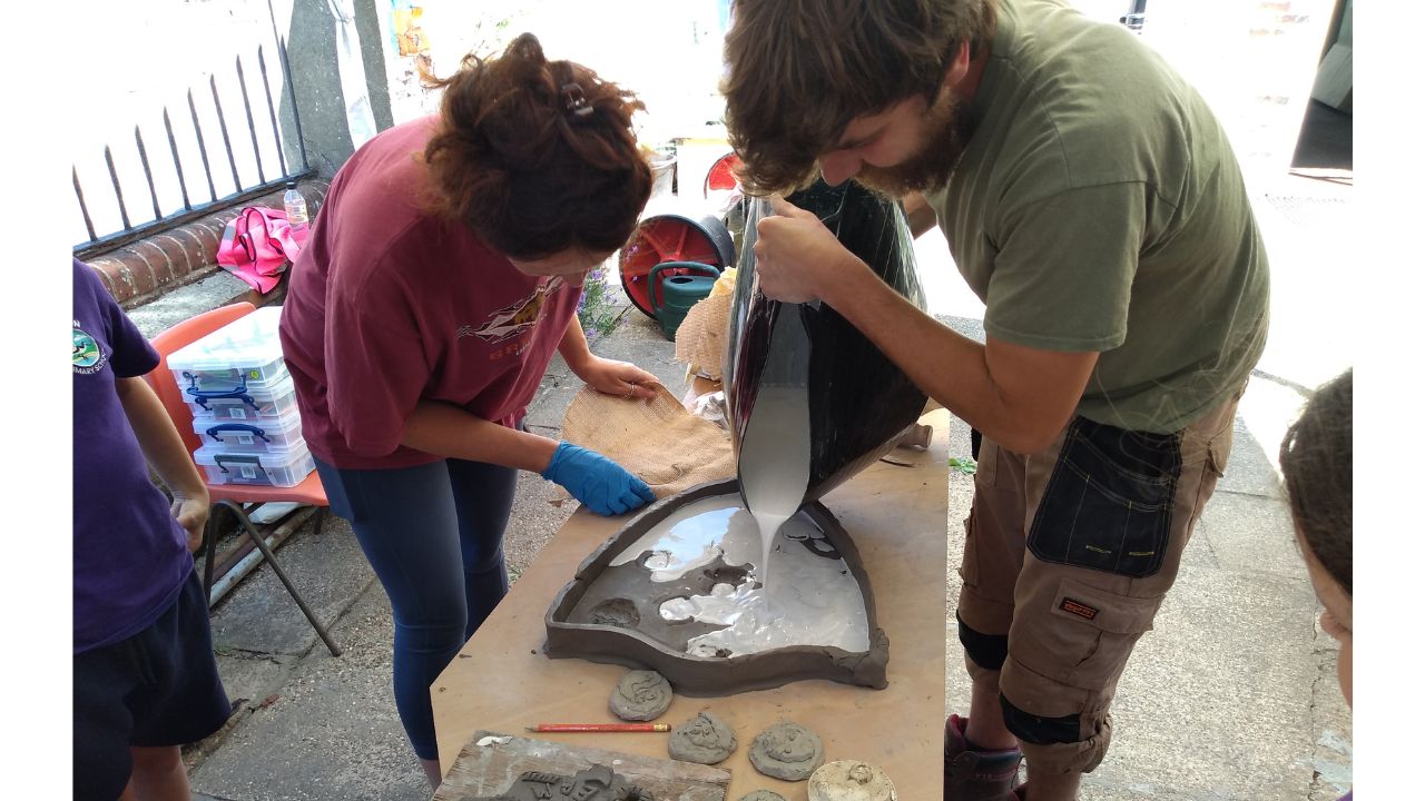 Two adults either side of a wooden table, carefully pour plaster into the clay mould