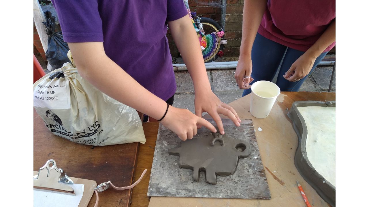 Two figures in the background, one obviously a child who is applying a clay shape to a slab of clay