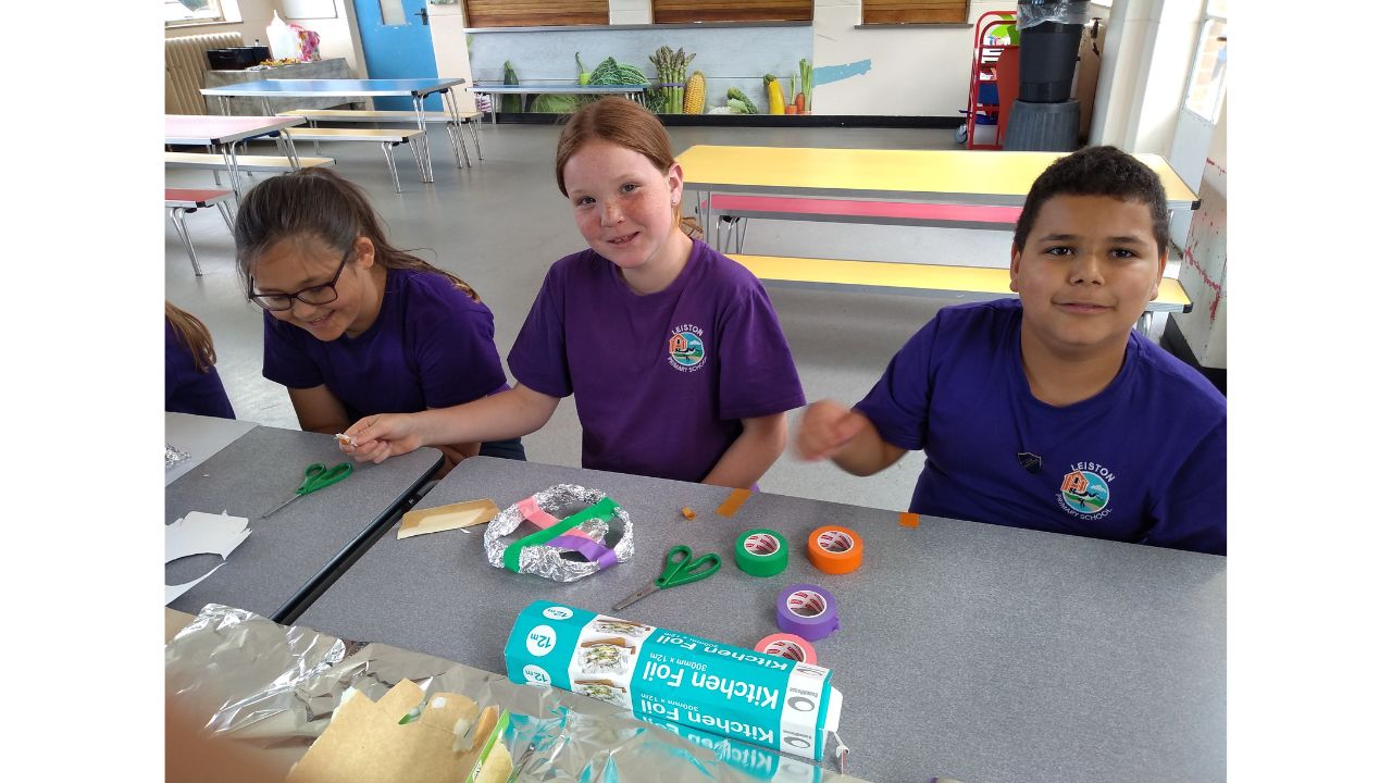 Three school children in purple t-shirts seated at a school dinner table with coloured tape, silver foil and other craft materials in front of them