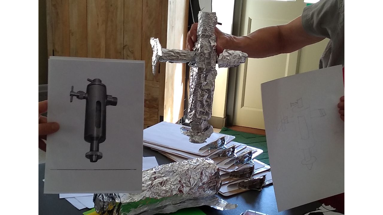 A person holds up a print of a precision engineered piece whilst a second person holds up a silver-foil covered model of the piece and a drawing of same