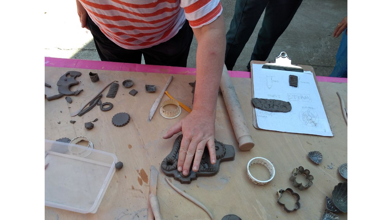 A person in an orange and white striped T shirt leans forward over a table on which are pieces of clay and assorted tools for working with clay. She is smoothing a piece of clay on to the surface of a larger piece