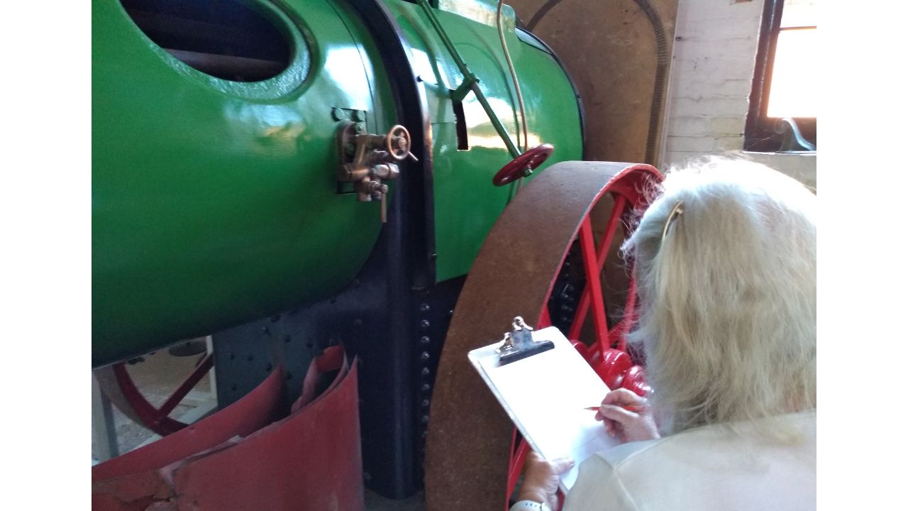 A woman stands in front of a large green and red steam engine, noting down something on her clip board