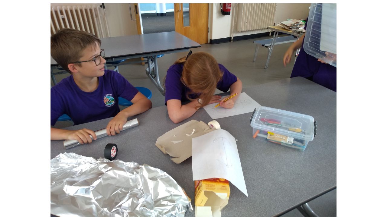 A child leans over a piece of paper, drawing, whilst another seated beside her holds a roll of silver foil. On the table in front of them are various craft materials