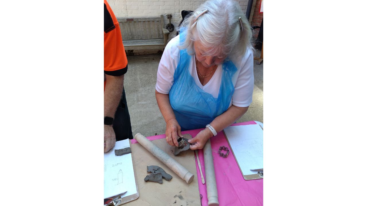 A woman wearing a white T shirt and blue plastic apron fashions a piece of clay in her hands whilst on the table in front are two rolling pins and a cutter