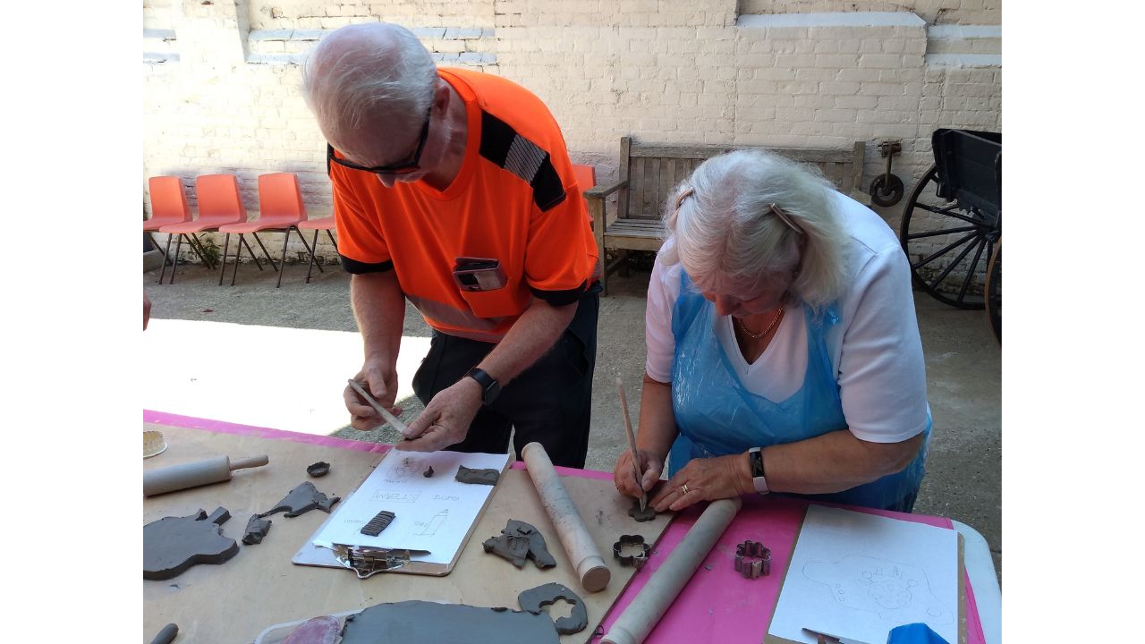 A man in an orange top stand beside a woman, both leaning over a table and using wooden tools to mark make in the clay. On the table in front of them are rolling pins, pieces of clay and cutters