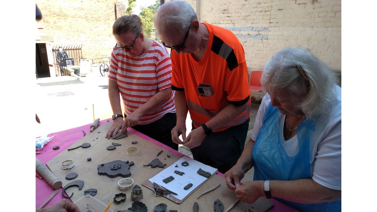 The people stand in front of a table on which are pieces of clay, cutters and rolling pins. They are all busy mark making
