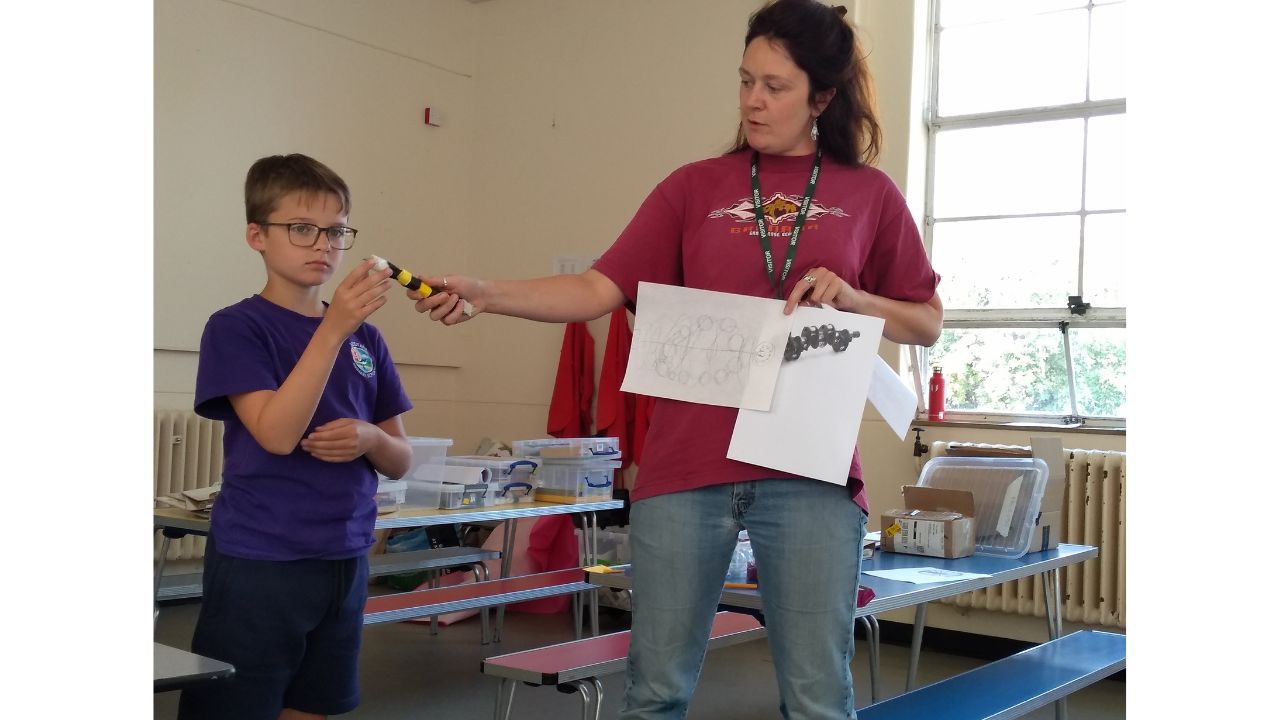 An adult stands holding several drawings whilst a child beside her holds up a black and yellow tube with moving parts