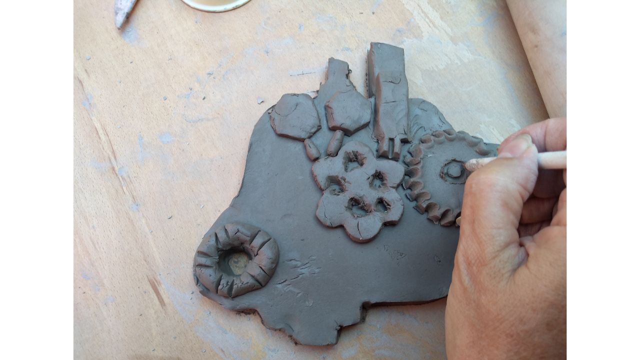 Overhead view of clay slab with smaller pieces of clay adorning it and a hand on the right using a wooden stick to make finer details