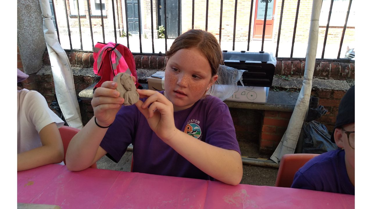 A child seated at a table with pink covering, holds up a clay disc on which is fashioned a face