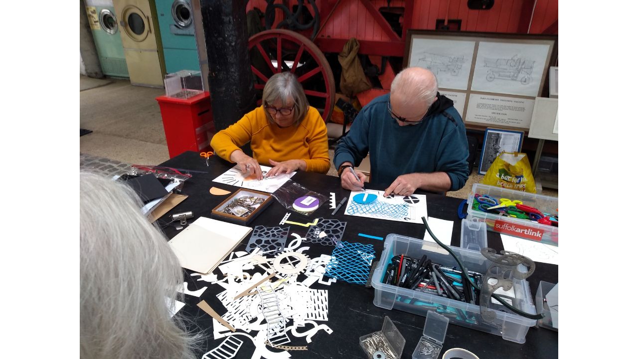 Two adults seated at a long table covered with drawing materials, creating individual images
