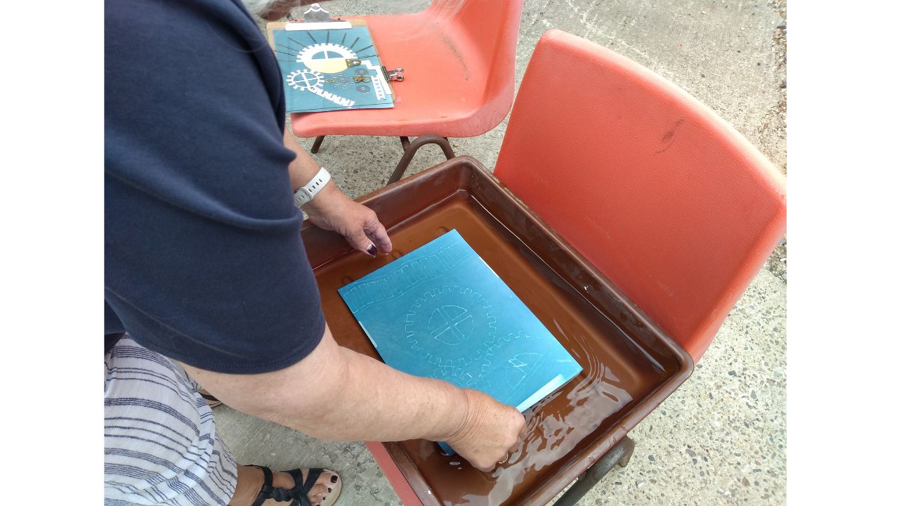 An overhead view of a tray of water on an orange chair and a person rinsing a blue picture in the water in the tray