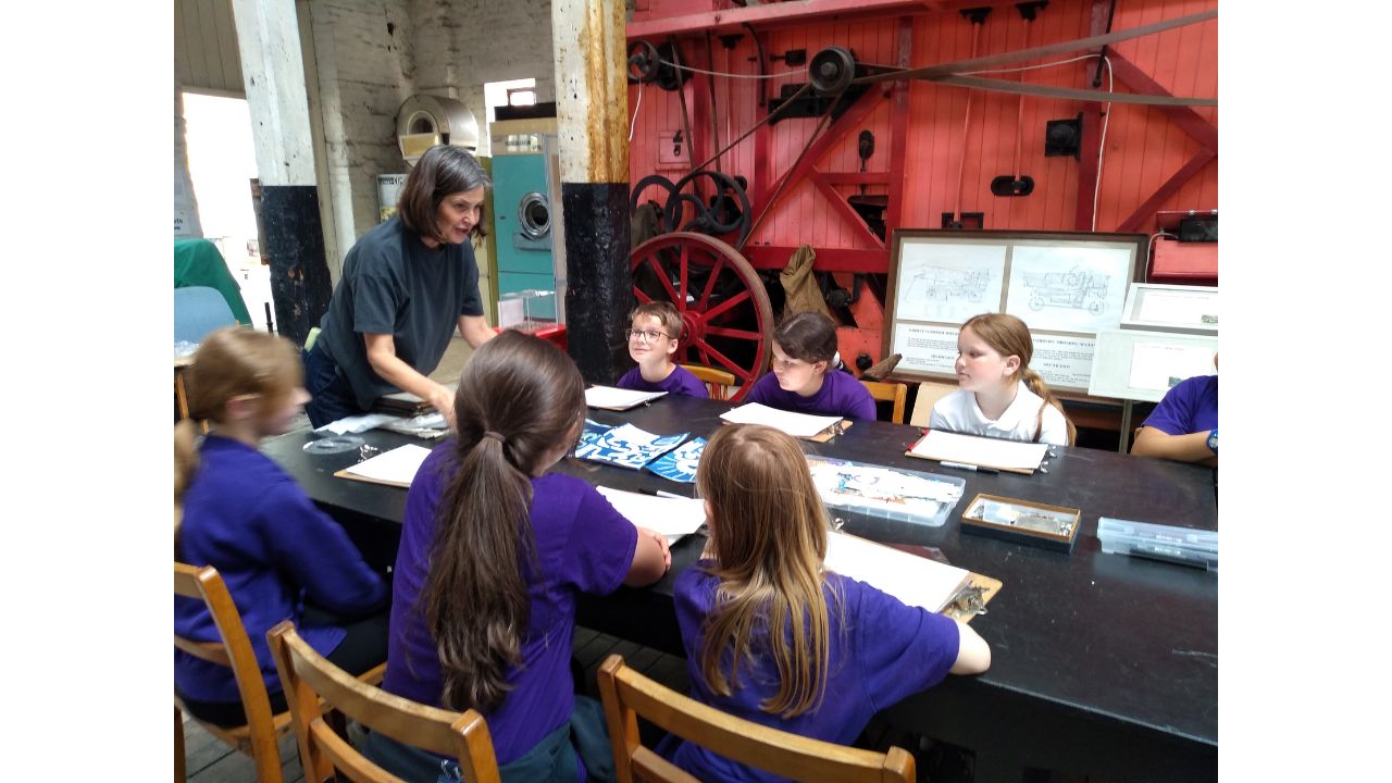 Two rows of children seated at a long dark table, with a huge red old fashioned thresher behind them