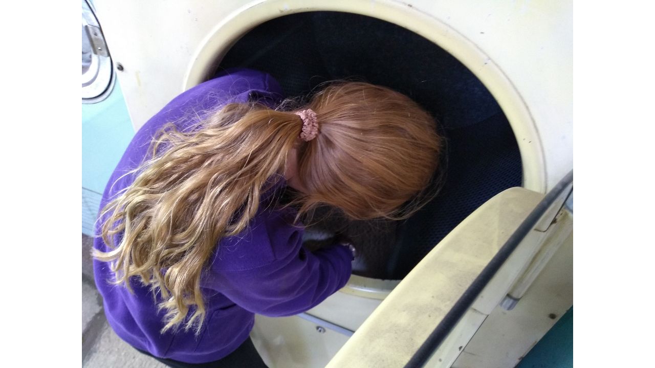 A child with long auburn hair tied back in a pink band is peering inside the large drum of an enormous industrial washing machine