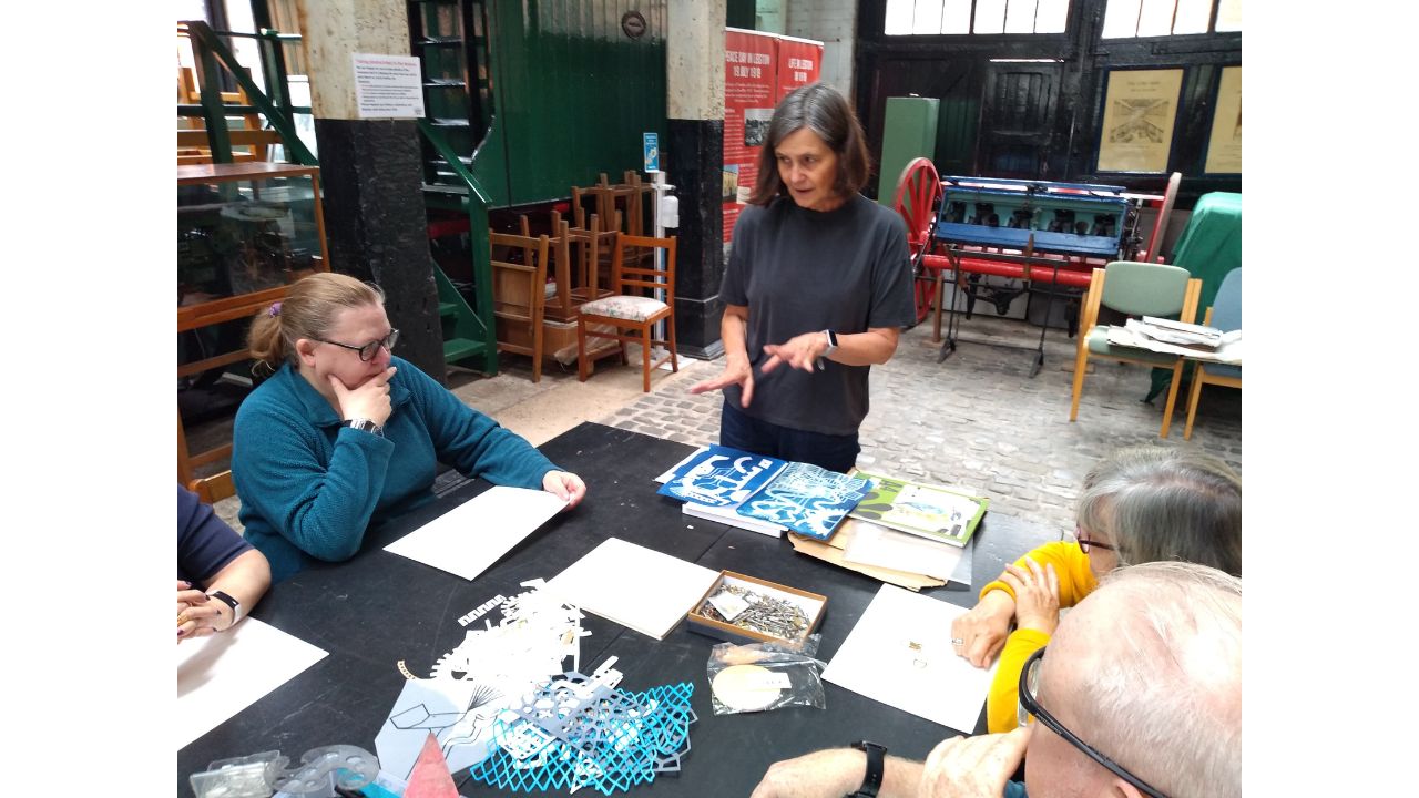 A woman in a dark T-shirt and trousers stands at the head of a table obviously discussing something with a group of adults, presumably about the pictures in front of her
