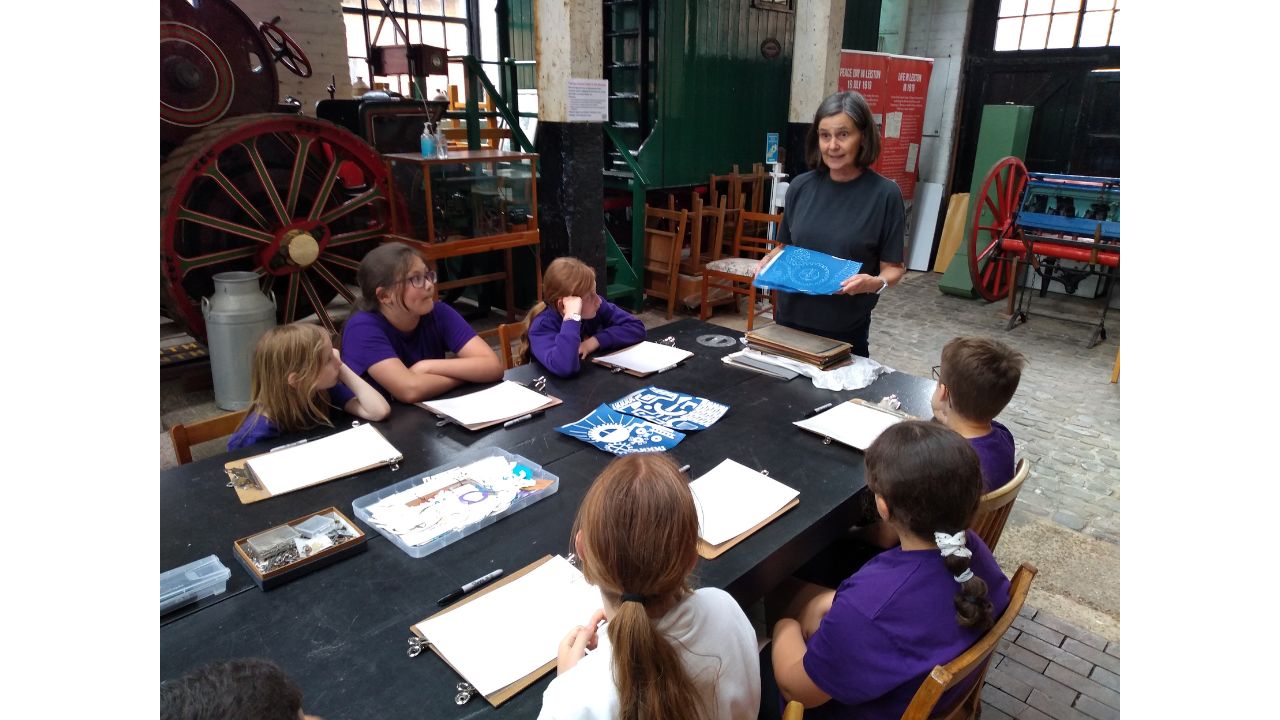 A woman in a dark T shirt and trousers stands at the head of a long dark table, holding up a cyanotype picture whilst children seated either side of the table watch her