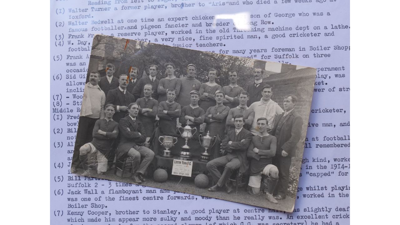 A photograph of Leiston Town FC in 1912-13 with three trophies and two footballs in the foreground and players and other members standing or seated in three rows. The photo is on a typewritten page with numbers down the side, each number referencing an individual player with name and a little bit of information about them