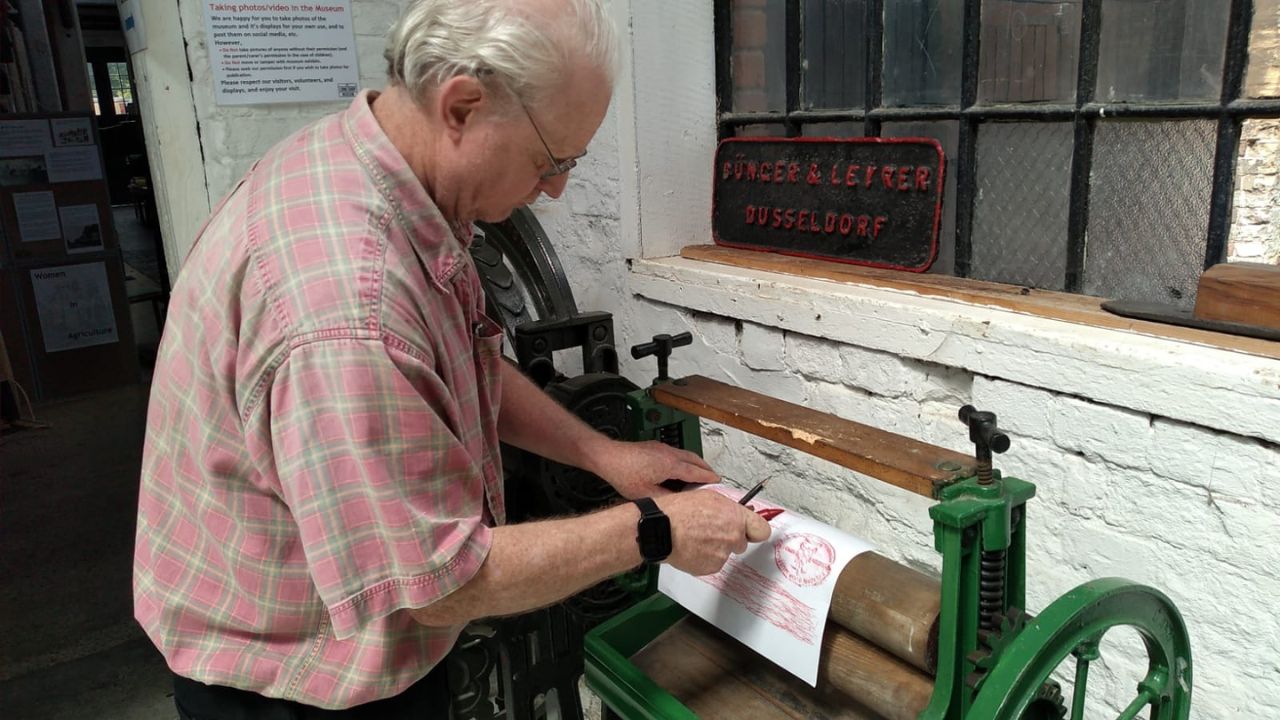 A gentleman stands in front of an old fashioned mangle and has draped a piece of paper over one of the wooden rollers, taking an impression of it by rubbing with a red crayon