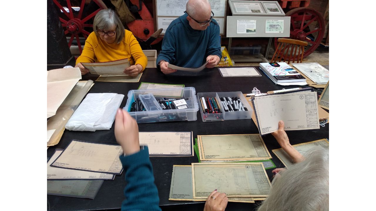 Adults seated around a table looking at an archive collection of technical drawings, tracings and blue prints