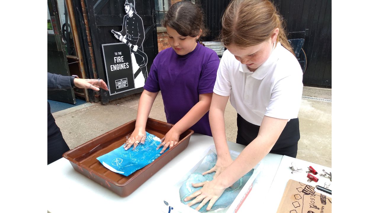 Two school girls stand in front of trays of water in which are cyanotype pictures