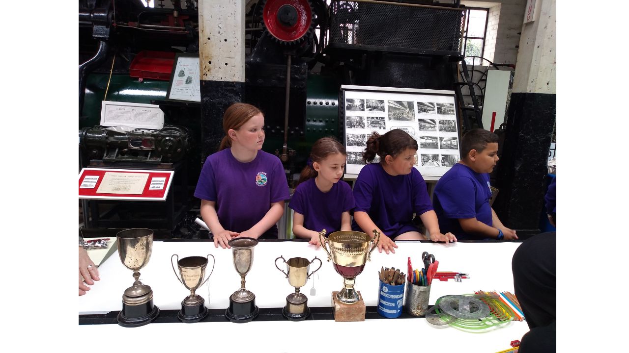 A row of 4 school children, each wearing a dark blue/purple T-shirt, and on the table in front of them is a row of 5 trophies plus pots of pencils and scissors