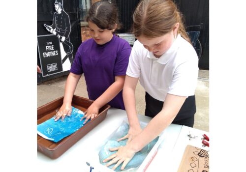Two school girls, one in a purple and the other in a white T shirt have submerged sheets of A4 paper into shallow trays of water and are washing them as the picture emerges in the light sensitive blue paper