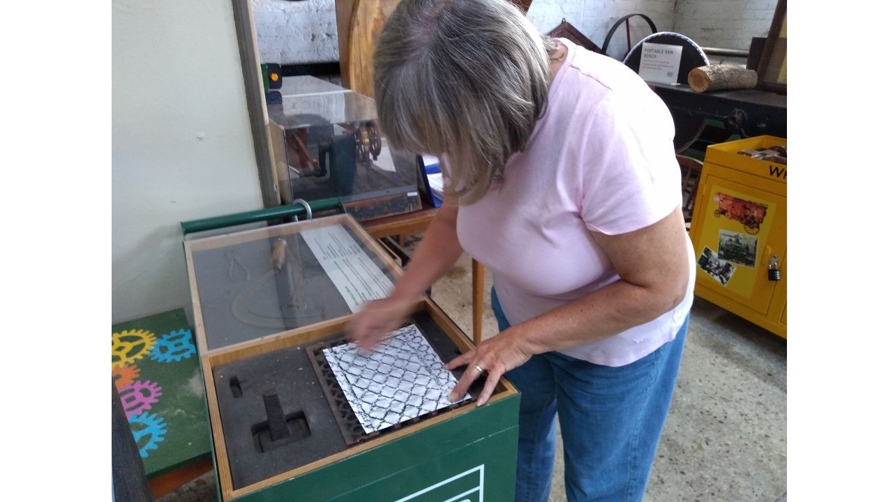 A woman in blue jeans and pink T-shirt leans over a piece of paper which is laid on top of an old iron grating - she is taking a rubbing of it using a graphite pencil