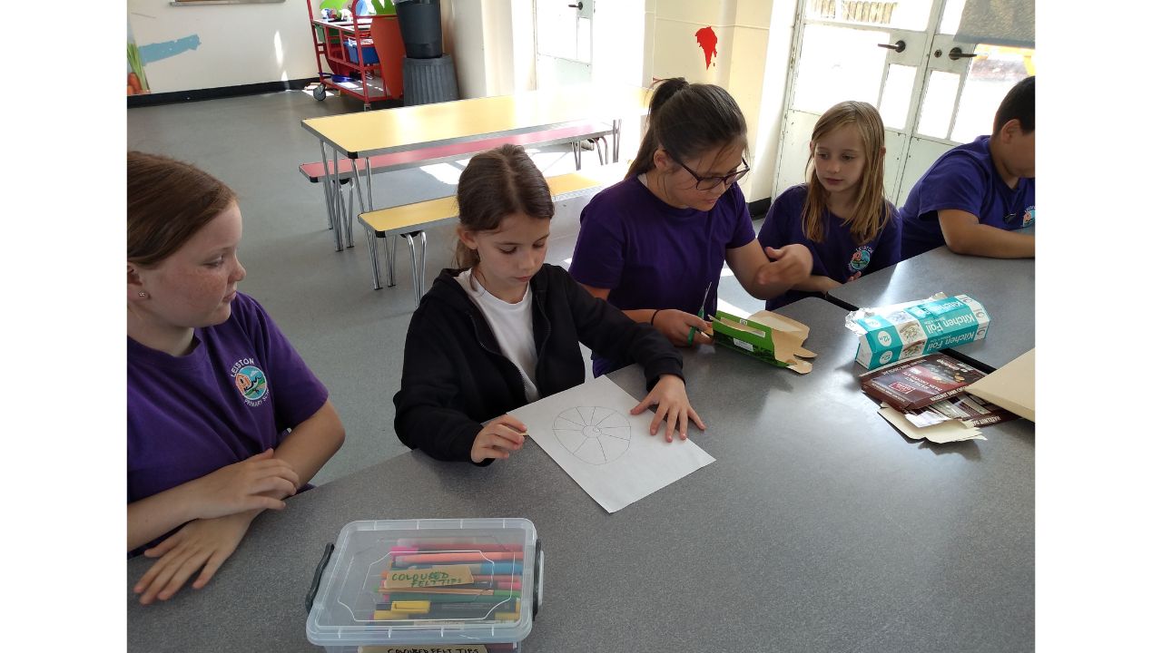 A row of school children seated at a long table. Pencils, paper and cardboard in front of them. One child studies a picture whilst another folds cardboard