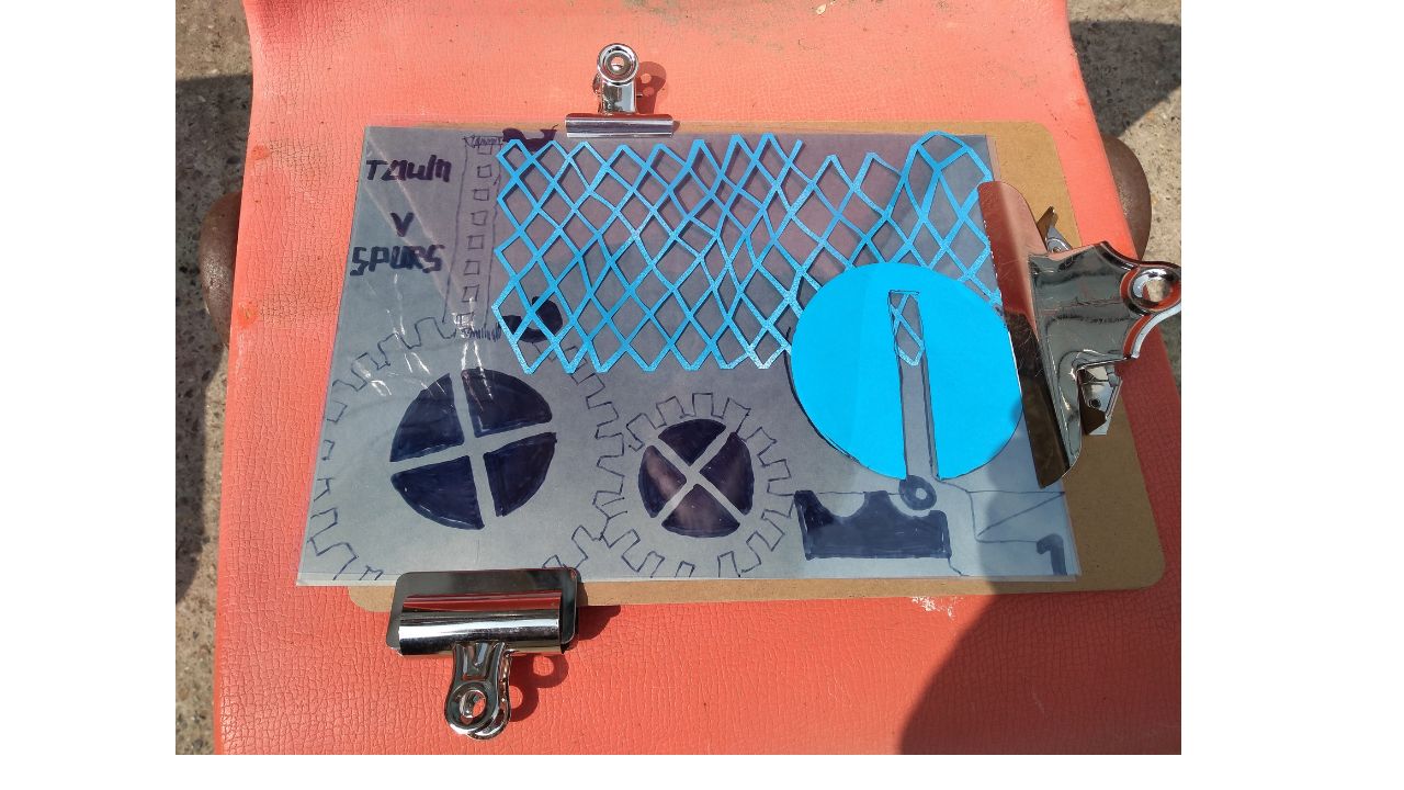 On a plastic orange chair, an A4 clipboard holds a piece of light sensitive paper, on which are paper shapes and drawings of cogs
