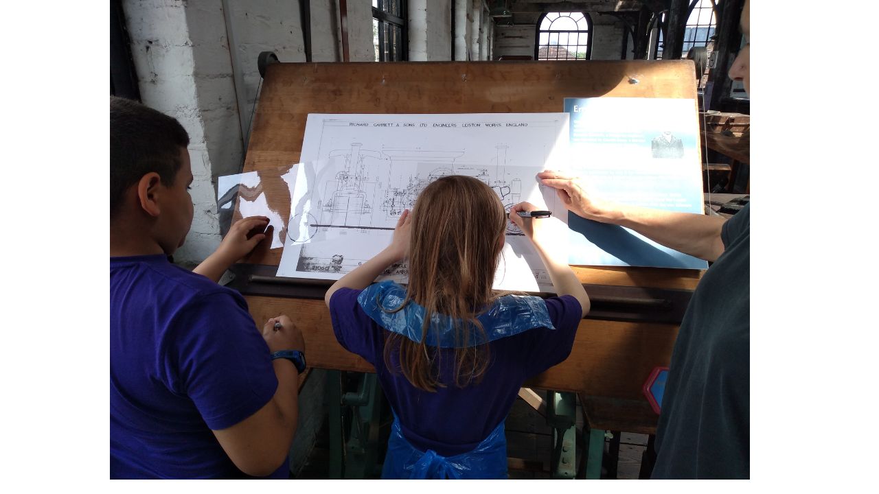 Two children stand at an old fashioned drawing board drawing over a technical picture as an adult stands to the right of picture