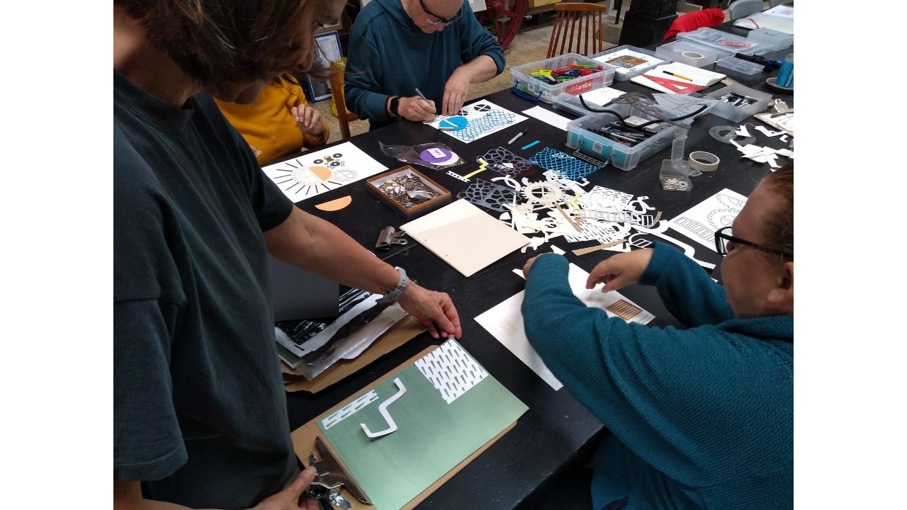 A table covered in different shapes, pens and other drawing materials, people drawing and arranging small paper shapes