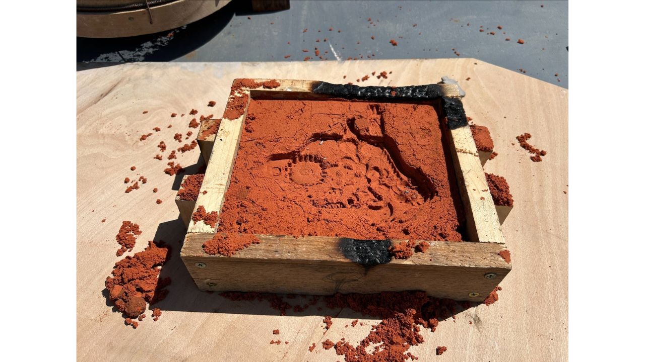 A wooden frame filled with red sand and an indentation in it