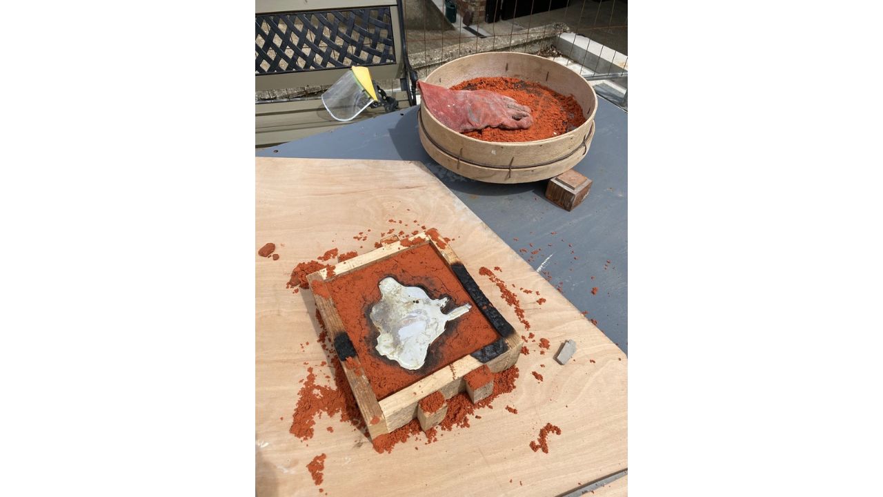 The sand mould filled with pewter with sieve and eye protector shield beside