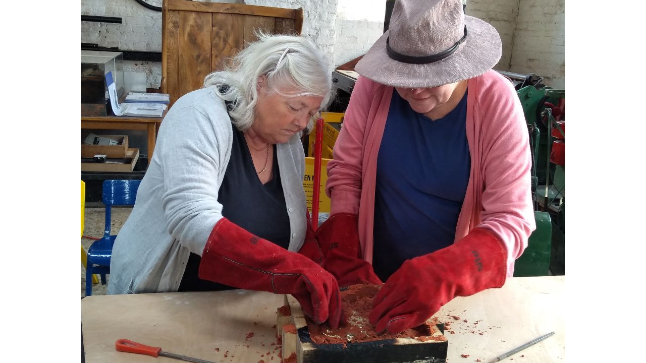Two women standing over wooden frame, each wearing long red leather gloves. One wears a hate with a dark band