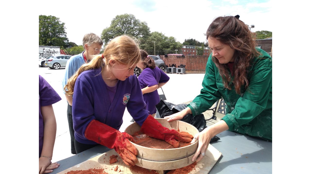 Woman in green overall holds large wooden sieve whilst child wearing huge red leather gloves pushes red sand through the mesh