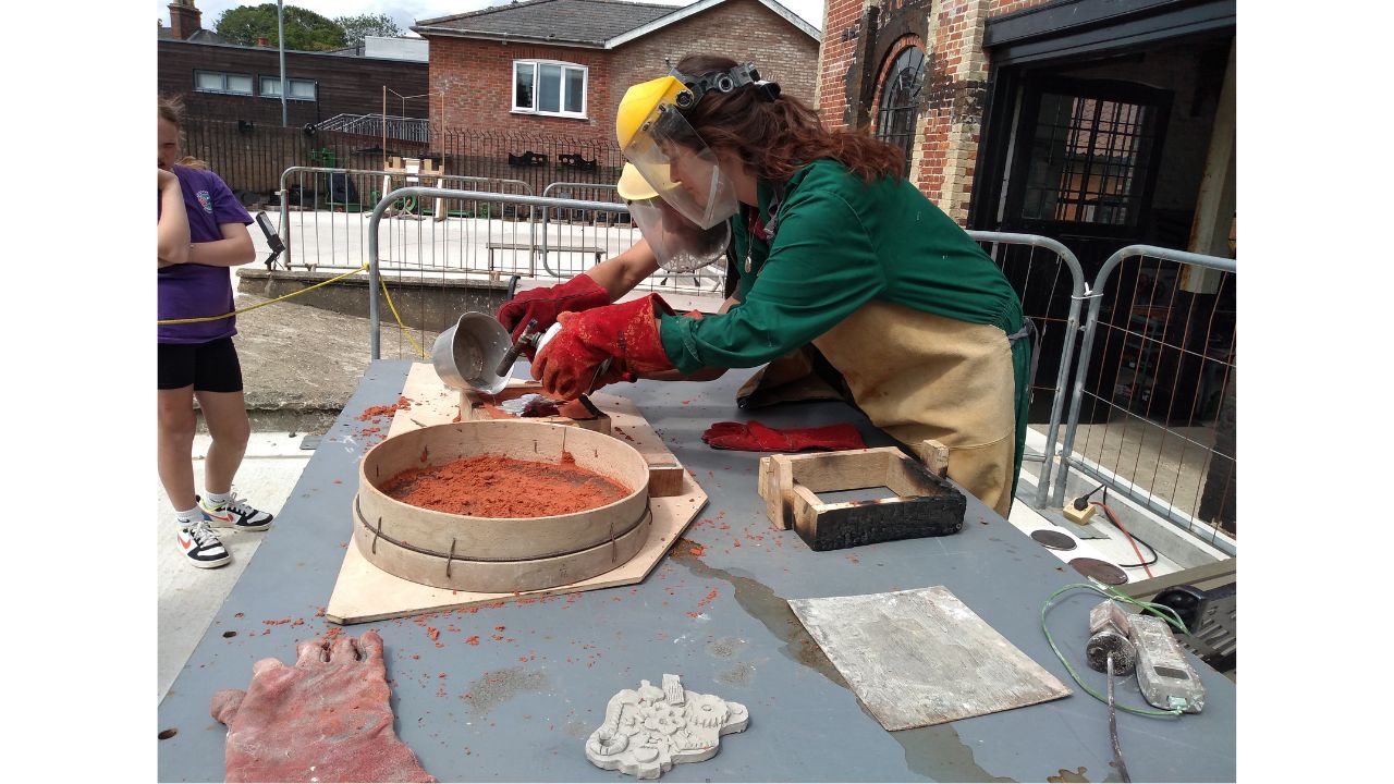 Woman in green overall and face protector leans over mould as a second adult pours from a small metal saucepan. Child looks on